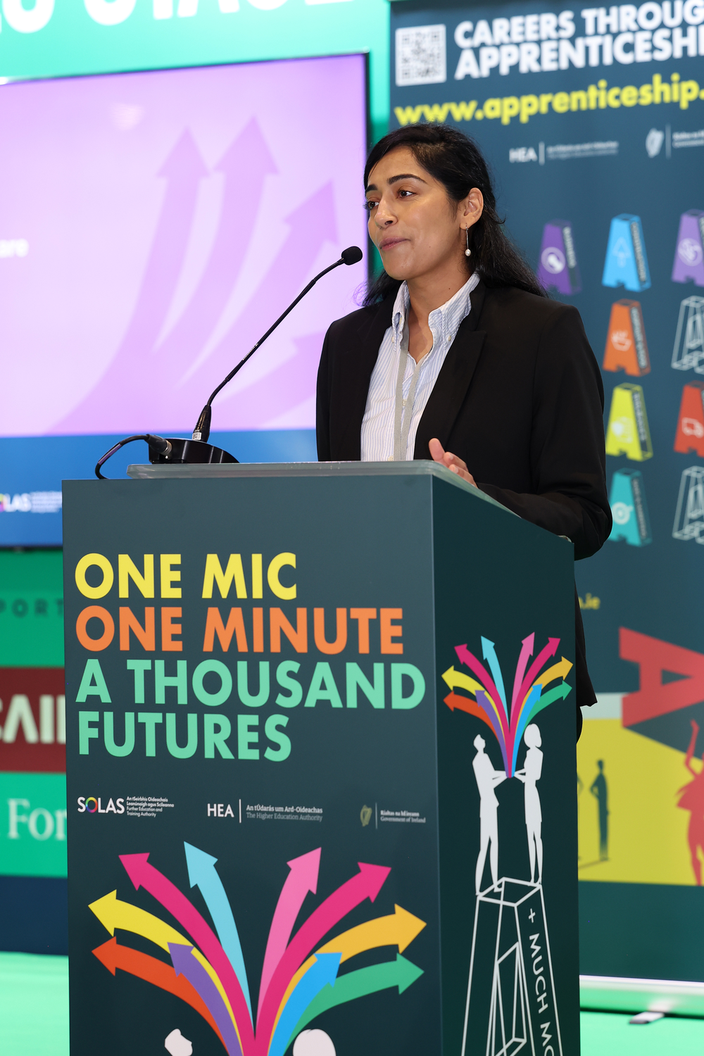 A woman in a suit speaks at a podium with the text 